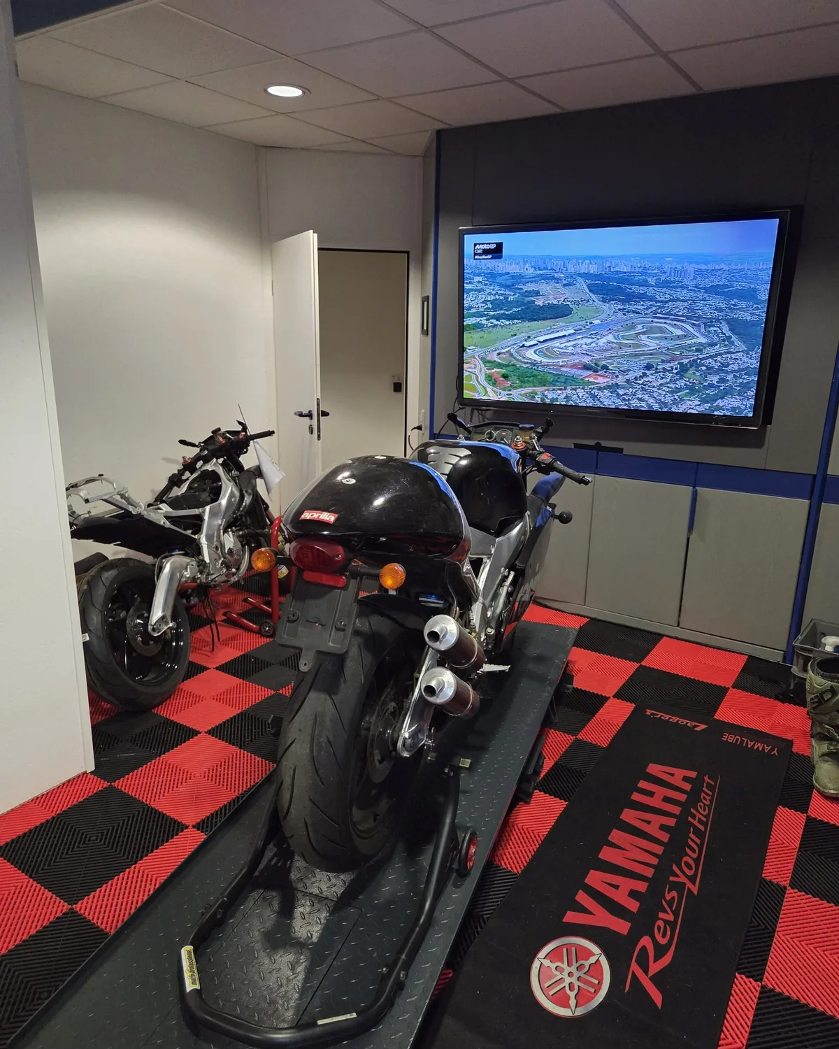 Werkstatt von Motorrad Friedrich: Aprilia auf der Hebebühne, dahinter ein zerlegtes Motorrad, an der Wand läuft MotoGP auf dem Bildschirm